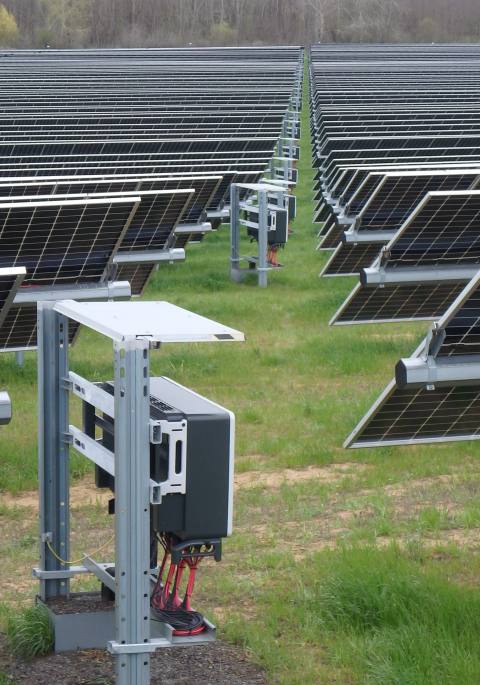 String-Wechselrichter an der Unterkonstruktion einer PV-Freiflächenanlage, dahinter Modulreihen