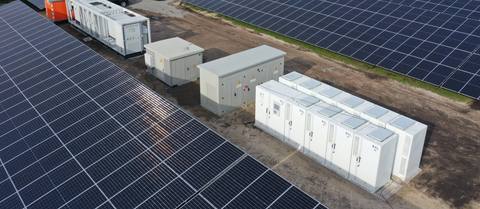 Luftaufnahme eines Batteriespeichers in einem Solarpark: weiße Speicherschränke in einer Reihe, daneben Stationsgebäude und ein Container, ringsum Modulreihen