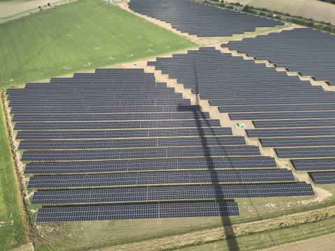 Luftbild einer Freiflächen-Photovoltaikanlage, über die Modulreihen fällt der Schatten einer Windenergieanlage