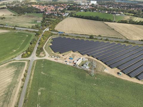 Aerial view of a ground-mounted photovoltaic installation between fields and farm tracks, with a village in the background