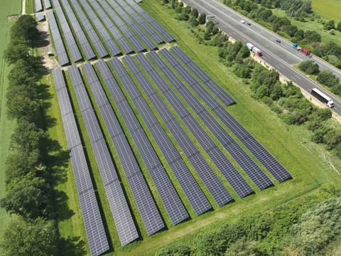 Aerial view of a ground-mounted photovoltaic system directly next to a motorway, with a strip of trees and shrubs in between