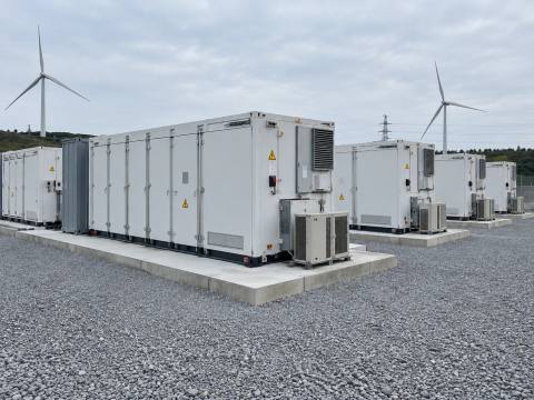 Batteriespeicher-Container mit Klimageräten auf Betonfundamenten, im Hintergrund Windenergieanlagen